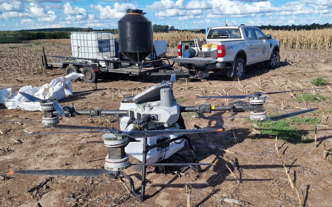 DJI Agras T100 von Kunz Agrotech auf einem abgeernteten Feld in Uruguay, dahinter das Begleitfahrzeug und ein Anhänger mit Wassertank und Mischtechnik.